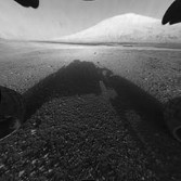 Curiosity sees two wheels on soil, its shadow in front of it, a dark line of sand dunes, and a bright hill of the Gale crater central mountain in this photo taken from its front hazard avoidance camera late on the day it landed.